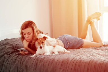 Köpeğiyle yatakta dinlenen ve akıllı telefon kullanan bir kadın.