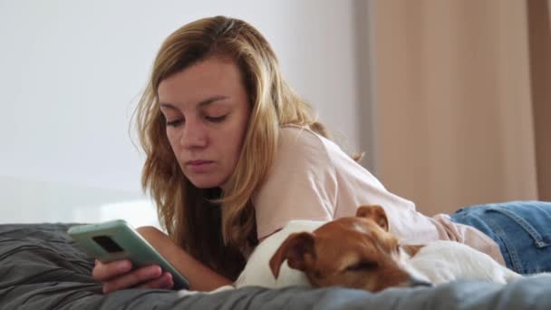 Mujer descansando en la cama con su perro y usando smartphone — Vídeo de stock