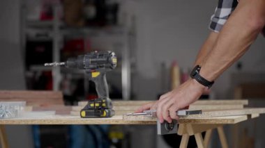 Close-up of carpenter drilling hole in wooden panel using drill and metal jig tool for precision. Man working in home workshop. Concept of woodworking, furniture making and DIY home projects