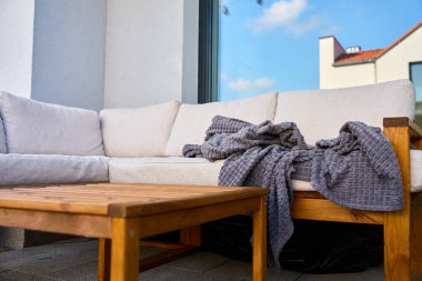 Açık köşedeki kanepe, hafif yastıklar, ahşap masa ve fayanslı terasta gri battaniye. Rahatlama, avlu konforu ve modern açık hava yaşam tarzı kavramı