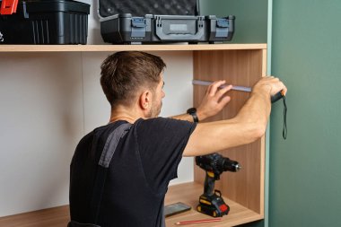 Evdeki mobilya montajı sırasında paneli işaretlemek için bant ve kalem kullanan adam ahşap dolabı ölçüyor.