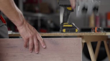 Close-up of carpenter drilling hole in wooden panel using drill and metal jig tool for precision. Man working in home workshop. Concept of woodworking, furniture making and DIY home projects