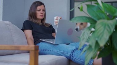 Terasta uzaktan çalışırken elinde laptop ve kahve bardağıyla kanepede oturan bir kadın. Uzaktan çalışma, ev ofisi, teknoloji ve modern yaşam tarzı kavramı