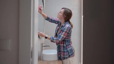 Modern banyoda lavabonun yanında pembe bezle ayna temizleyen gözlüklü bir kadın. Ev temizliği, hijyen, günlük rutin ve ev işleri kavramı