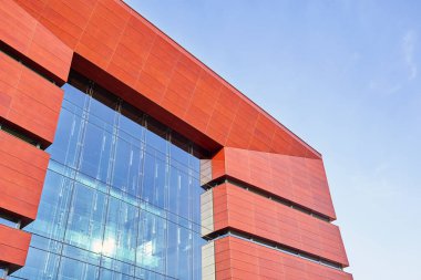 Kırmızı metal panelleri ve mavi gökyüzünü yansıtan büyük camlı modern binanın düşük açılı görüntüsü. Çağdaş mimari ayrıntıları