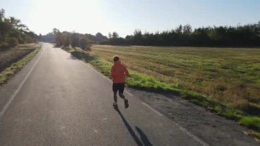 Gün doğumunda boş kırsal yolda turuncu ceketli ve şortlu koşan bir adamın arka görüntüsü. Sabah koşan adam. Zindelik ve sağlıklı yaşam tarzı kavramı