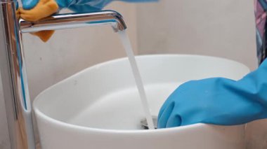 Mavi eldiven giyen bir kadın banyo musluğunu temizliyor ve beyaz lavaboyu sünger ve deterjanla temizliyor. Hijyen, temizlik, ev işleri ve günlük bakım kavramı