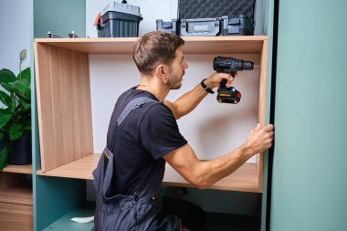 Tahta dolapta elektrikli matkapla delik açan bir adam. Dairenin yenilenmesi sırasında ev mobilyası montajı