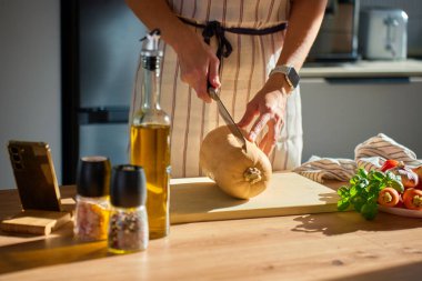Mutfaktaki tahta tahtada tereyağlı kabak kesen önlüklü bir adam. Masada taze sebze ve otlarla taze balkabağı dilimleyen bir kadının yakın çekimi. Sağlıklı beslenme ve ev yemeği kavramı