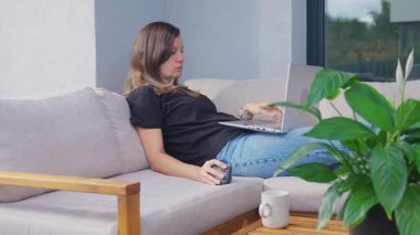 Terasta dizüstü bilgisayarla çalışan serbest çalışan bir kadın. Kanepede dinleniyor. Uzaktan çalışma, konfor ve dijital yaşam tarzı dengesi kavramı