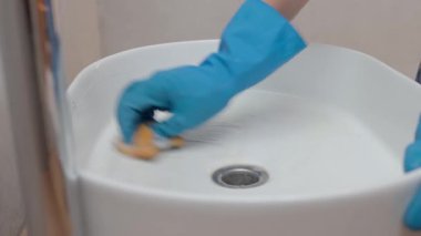 Banyoda beyaz seramik lavaboyu sünger ve köpükle temizleyen mavi eldivenli bir kadın. Hijyen, temizlik, ev işleri ve günlük bakım kavramı