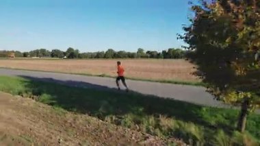 Güneşli bir günde tarla ve yeşil ağaçların yanında boş kır yolunda koşan bir adam. Atletizm açık havada. Sabah koşusu. Spor anlayışı, açık hava aktivitesi ve sağlıklı yaşam tarzı.