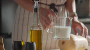 Bir kadın mutfak masasına kremalı ölçü kabını yerleştirirken yemek hazırlıyor. Ev mutfağında süt, zeytinyağı, tuz, biber ve kabakla pişirme işlemi