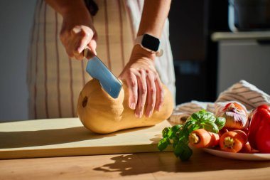 Mutfaktaki tahta tahtada tereyağlı kabak kesen önlüklü bir adam. Masada taze sebze ve otlarla taze balkabağı dilimleyen bir kadının yakın çekimi. Sağlıklı beslenme ve ev yemeği kavramı