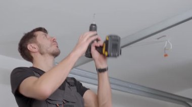 İş tulumu giyen bir adam tavan arasında kablosuz matkapla delik açıyor. Ev yenileme, kurulum ve profesyonel bakım hizmeti kavramı.