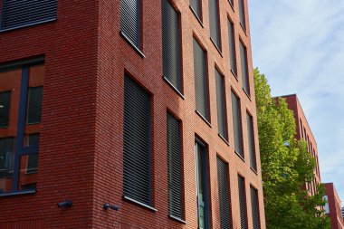 Gündüz vakti dikey pencere panjurları ve ağaç olan modern kırmızı tuğla bina cephesi. Mimarlık, dış ve şehir gelişimi kavramı.