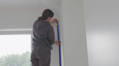 Mavi maskeleme bandını evin içinde dikey duvar köşesine yapıştıran kadın. İç yenileme, duvar boyama hazırlığı, ev onarımı ve DIY bitirme işi kavramı.