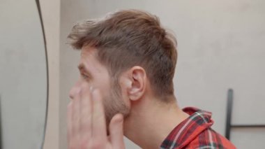 Modern banyoda aynaya bakarken kırmızı pijamalı bir adam yüz kremi sürüyor. Erkek derisi, bakımı, kendine bakımı ve günlük hijyen kavramı