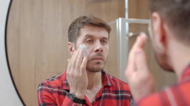 Adamın banyoda yüzüne beyaz maske kremi sürerken yakın plan fotoğrafı. Erkek derisi, yüz bakımı ve kişisel hijyen kavramı