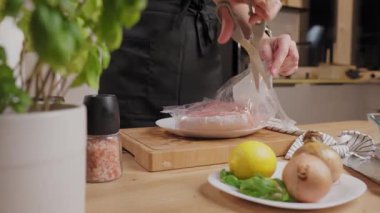 Ev mutfağındaki bir tabakta çiğ tavuk filetosu ile plastik ambalajı kesen kadın elleri. Yemek hazırlama ve ev yemeği kavramı