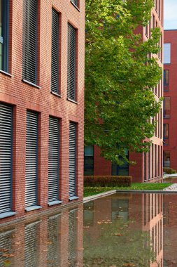 Pencere panjurları ve sığ sulara yansıyan ağaçlı modern kırmızı tuğla bina. Mimarlık, kentsel tasarım ve dış detay kavramı.