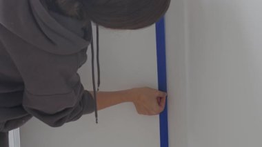 Mavi maskeleme bandını evin içinde dikey duvar köşesine yapıştıran kadın. İç yenileme, duvar boyama hazırlığı, ev onarımı ve DIY bitirme işi kavramı.
