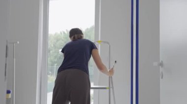 Modern odadaki pencerenin yanındaki duvarı fırçayla boyamak için merdiven kullanan kadın. Ev yenileme, iç boyama ve DIY onarım kavramı.