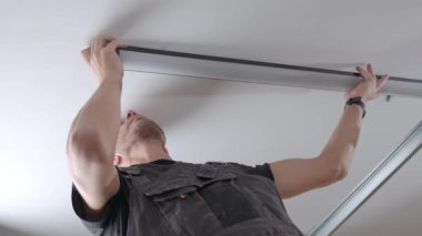 İş kıyafetli bir adam evin tavanına doğrusal LED lamba takıyor. Elektrik tesisatı, bakım işleri ve ev yenileme kavramı.
