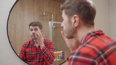 Modern banyoda aynaya bakarken kırmızı pijamalı bir adam yüz kremi sürüyor. Erkek derisi, bakımı, kendine bakımı ve günlük hijyen kavramı