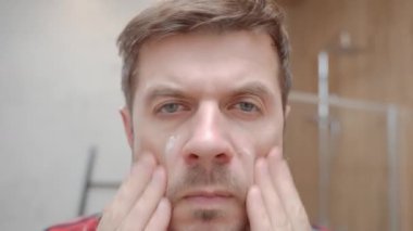 Modern banyoda aynaya bakarken kırmızı pijamalı bir adam yüz kremi sürüyor. Erkek derisi, bakımı, kendine bakımı ve günlük hijyen kavramı