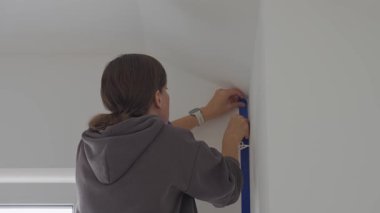 Mavi maskeleme bandını evin içinde dikey duvar köşesine yapıştıran kadın. İç yenileme, duvar boyama hazırlığı, ev onarımı ve DIY bitirme işi kavramı.