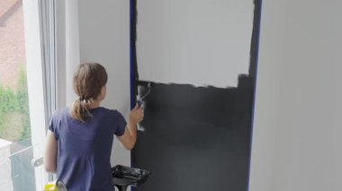Ev tadilatı sırasında, beyaz iç duvarı siyah renge boyayan bir kadın ve oturma odasında boya silindiri. DIY geliştirme ve apartman yenileme kavramı