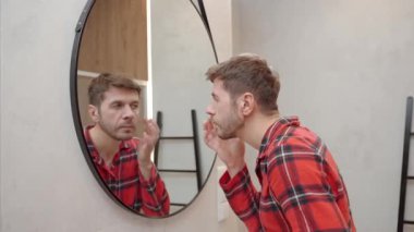 Modern banyoda aynaya bakarken kırmızı pijamalı bir adam yüz kremi sürüyor. Erkek derisi, bakımı, kendine bakımı ve günlük hijyen kavramı