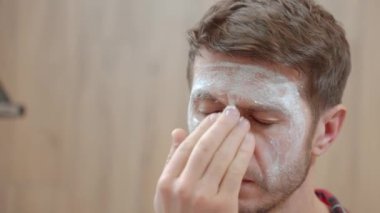 Adamın banyoda yüzüne beyaz maske kremi sürerken yakın plan fotoğrafı. Erkek derisi, yüz bakımı ve kişisel hijyen kavramı