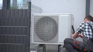 Dış binanın yakınındaki ısı pompası ünitesinde teknisyen bakım yapıyor. Adam aletleri kullanarak hava kaynağı ısı pompası kuruyor. HVAC hizmeti ve ısıtma sistemi onarımı kavramı