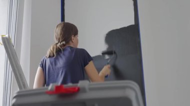 Ev tadilatı sırasında, beyaz iç duvarı siyah renge boyayan bir kadın ve oturma odasında boya silindiri. DIY geliştirme ve apartman yenileme kavramı