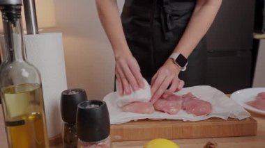 Kadın elleri ev mutfağında kağıt havluyla çiğ tavuk göğsü kurutuyor. Siyah önlüklü kadın taze sebze ve malzemelerle et filetosu hazırlıyor. Yemek pişirme ve yemek hazırlama süreci