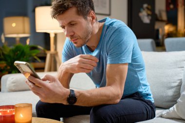 Oturma odasında kanepede oturan ve akıllı telefon kullanan bir adam. Evde cep telefonuyla dinlenen biri var. Çevrimiçi aktivite kavramı ve hayatta teknoloji kullanımı
