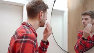 Banyoda aynaya bakarken yüzü pamukla temizleyen adam. Erkek derisi, yüz temizleme, günlük bakım ve kişisel hijyen kavramı..