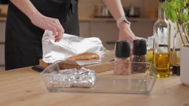 Kadın eli, baharatlı çiğ tavuk göğsünü alüminyum folyoyla mutfakta tahta kesme tahtasına sarıyordu. Yemek hazırlama ve pişirme süreci kavramı