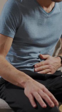 Oturma odasında koltukta oturan bir adam karın ağrısı ve kramplardan muzdarip. Sağlık sorunları olan kişi, sindirim sorunları veya evde gıda zehirlenmesi