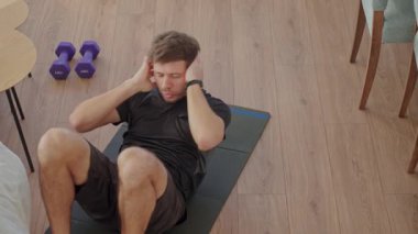 Atletik giysili bir adam evde, oturma odasında karın çatırdatma egzersizi yapıyor. Evde çalışma, temel eğitim, karın egzersizi ve sağlıklı yaşam tarzı kavramı