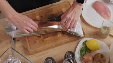 Kadın ellerinin baharatlı çiğ tavuk göğsünü alüminyum folyoyla mutfak tahtasına sarışının en üst görüntüsü. Yemek hazırlama ve pişirme süreci kavramı