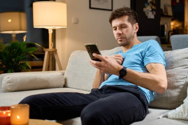 Oturma odasında kanepede oturan ve akıllı telefon kullanan bir adam. Evde cep telefonuyla dinlenen biri var. Çevrimiçi aktivite kavramı ve hayatta teknoloji kullanımı