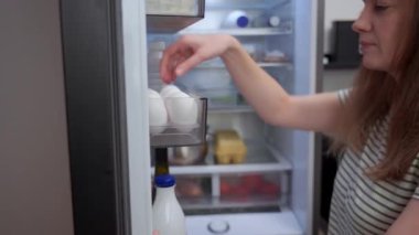 Kadın buzdolabını açıyor ve içinde yiyecek ve içecekler olan taze organik yumurtayı kapı rafından alıyor. Ev yemeği ve yiyecek depolama kavramı