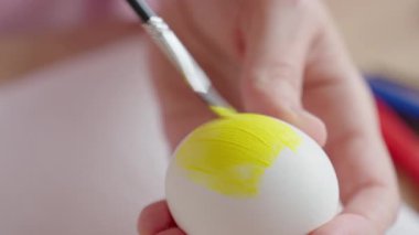Eller beyaz yumurtayı sarı boyayla boyuyor. Masadaki kağıt havlunun üzerinde küçük bir fırça kullanıyor. Paskalya yumurtası süslemesi ve ev yapımı tatil hazırlığı.