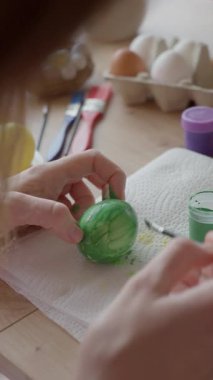 Beyaz yumurtayı fırça ve pastel boyayla boyayan kadın diğer renkli yumurtalar ve boya kavanozlarıyla aynı masaya oturuyor. Paskalya hazırlığı, yaratıcı hobi ve mevsimlik gelenek kavramı