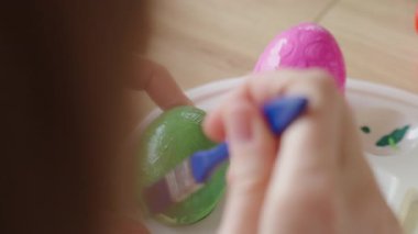 Masada fırçayla birlikte boya kavanozlarıyla beyaz yumurtayı yeşil boyayla boyayan ellerinizi kapatın. Paskalya yumurtalarını evde süsleyen kadın, tatile hazırlanıyor.