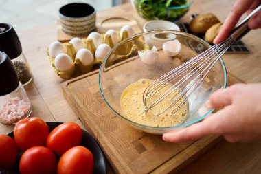 Ahşap mutfak tezgahında çırpılmış yumurtaları cam kasede çırpan kişi. Sağlıklı kahvaltı hazırlığı ve taze malzeme pişirme kavramı