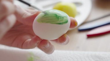 Masada fırçayla birlikte boya kavanozlarıyla beyaz yumurtayı yeşil boyayla boyayan ellerinizi kapatın. Paskalya yumurtalarını evde süsleyen kadın, tatile hazırlanıyor.